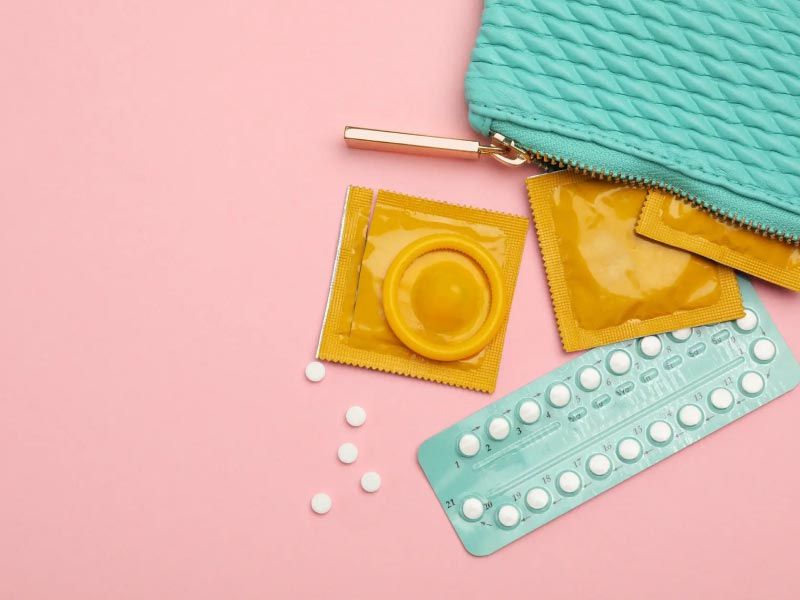 A green pouch with condoms and birth control pills spilling out on a pink surface