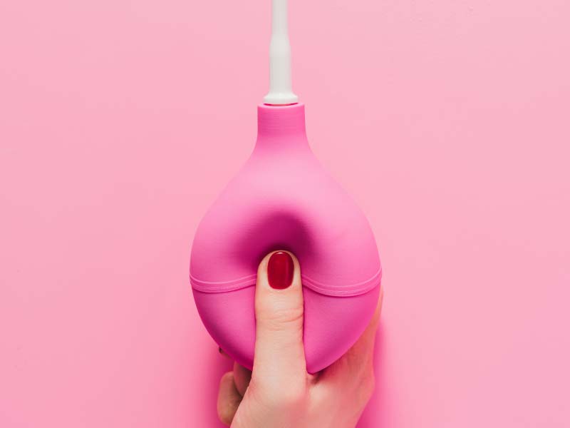 A woman's hand with red fingernails holding a pink vaginal douche bulb over a pink background