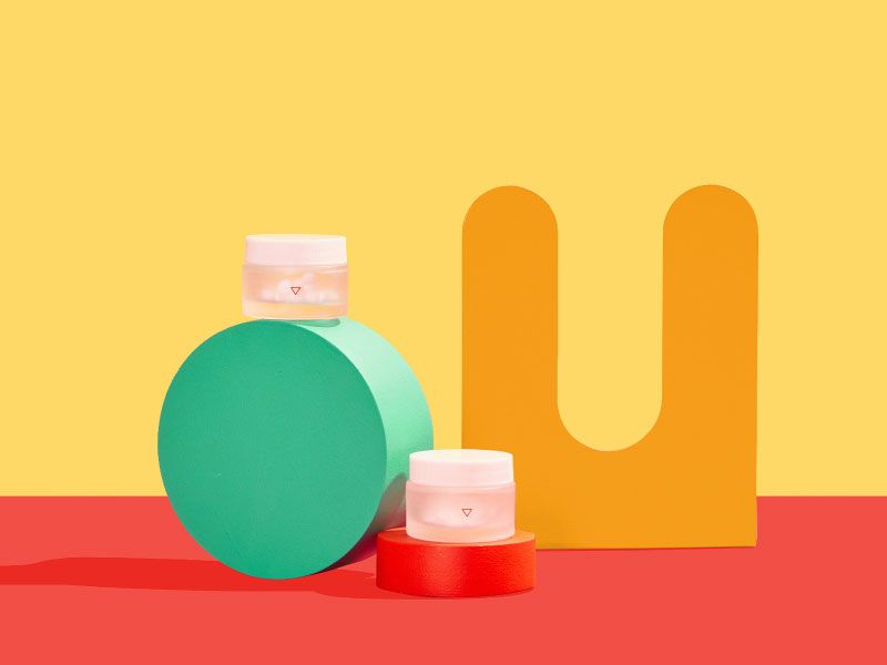 Two small Wisp glass pill jars balanced on colorful abstract shapes on a red surface with a yellow background