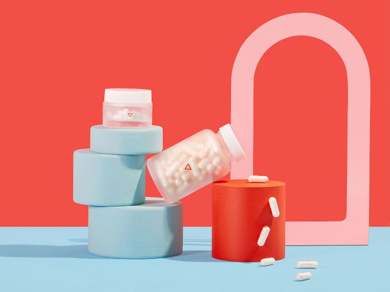 Two Wisp glass jars balancing on colorful abstract shapes will pills spilling out on a red and blue background