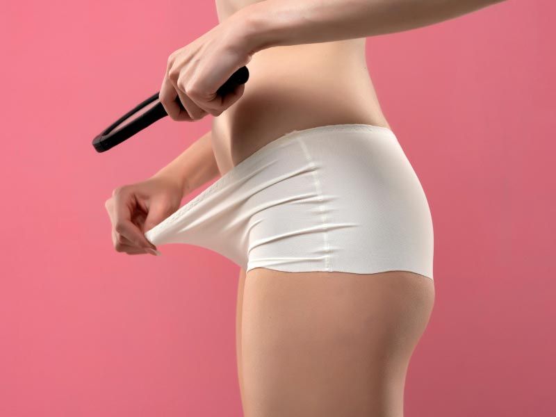 A woman standing with a magnifying glass looking inside her underwear over a rose-colored background