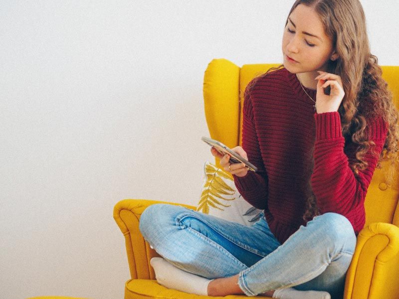 A female wearing a maroon sweater and blue jeans looking up information on her phone while sitting on a yellow chair
