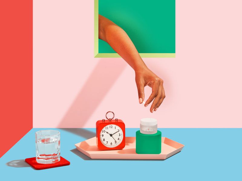 Woman's hand reaching for Norethindrone Acetate pills with an alarm clock on a pink tray and a glass of water on a red, pink and blue background