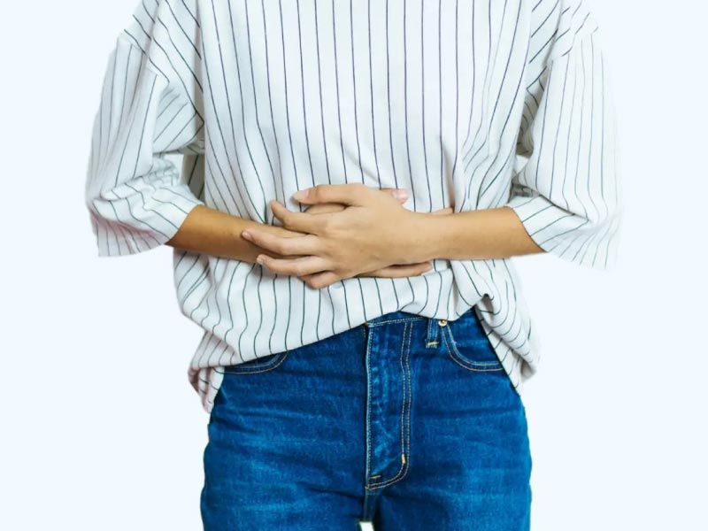 A woman wearing a striped white and black shirt with blue jeans has her hands over her abdomen in obvious discomfort
