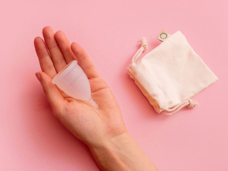 A woman's hand holding a menstrual cup with a storage bag next to it on a pink surface