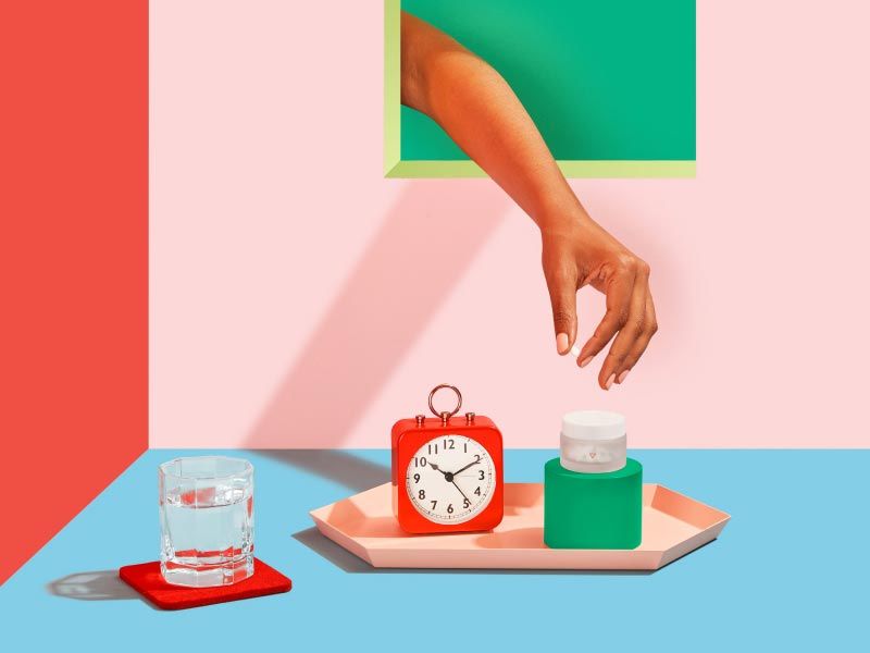 Wisp Delay Your Period medication with a red alarm clock, a glass of water and colorful abstract shapes on a red, pink, and light blue background