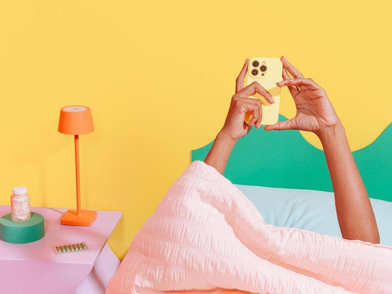 Female in bed ordering medication on her phone with a pink nightstand, orange lamp and Wisp glass medication jar with an orange and yellow background