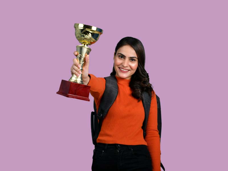 A woman wearing a red sweater, black pants and a black backpack holding a gold trophy in the air in front of a purple background