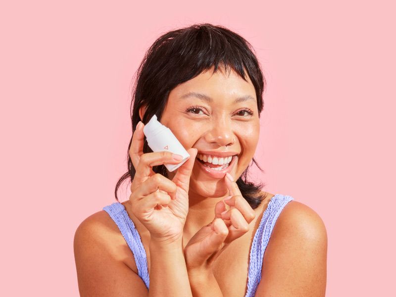 a smiling woman holding a bottle of Wisp herpes medication and applying it to her lip against a pink background