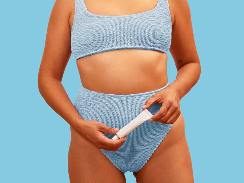 A woman wearing a blue bra and underwear is holding a tube of vaginal moisturizer