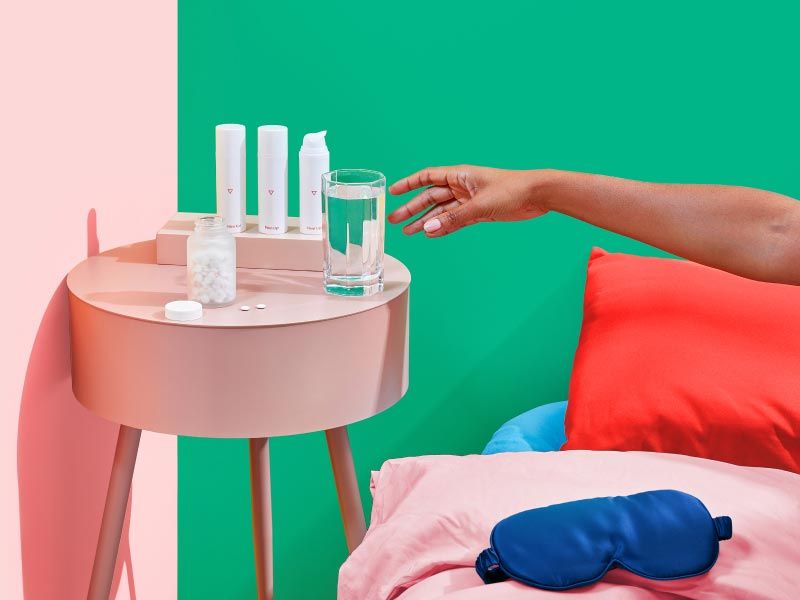 Woman's hand reaching for a glass of water on a pink nightstand with 3 bottles of Wisp Skincare treatment and a Wisp glass pill jar with pink and red bed and green and pink walls