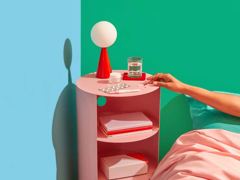 Woman's hand reaching for Wisp Telehealth Medication Abortion pills on a pink nightstand with a glass of water and a red lamp with blue and green walls and a pink comforter