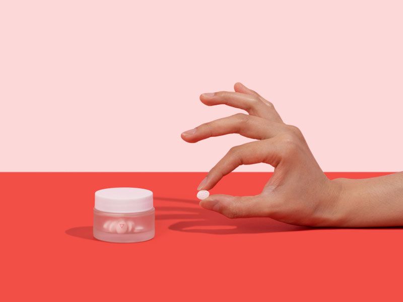 A woman's hand holding a Fluconazole pill next to a Wisp glass jar on a pink and red background