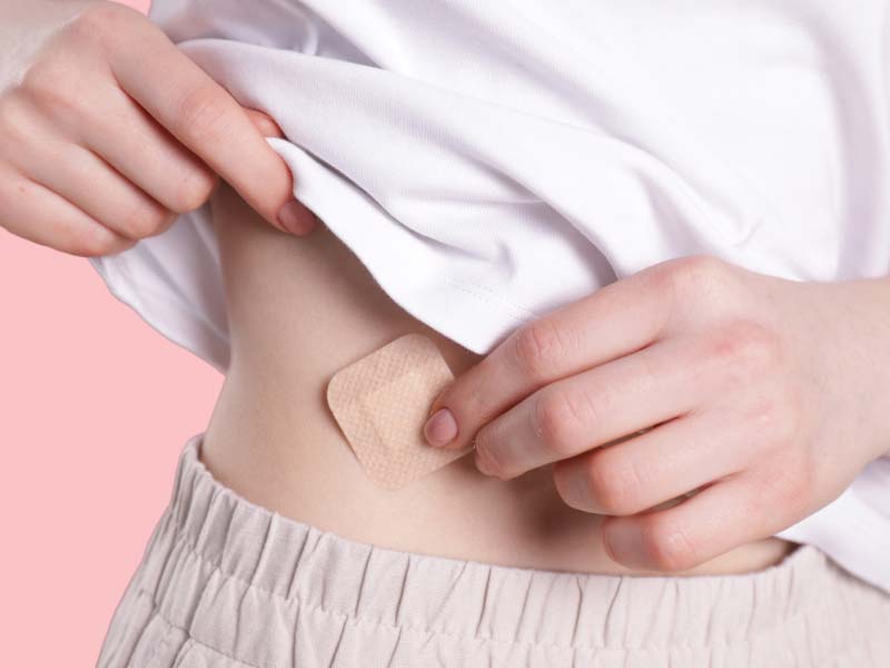 A woman wearing a white shirt and cream pants applying the birth control patch to her abdomen