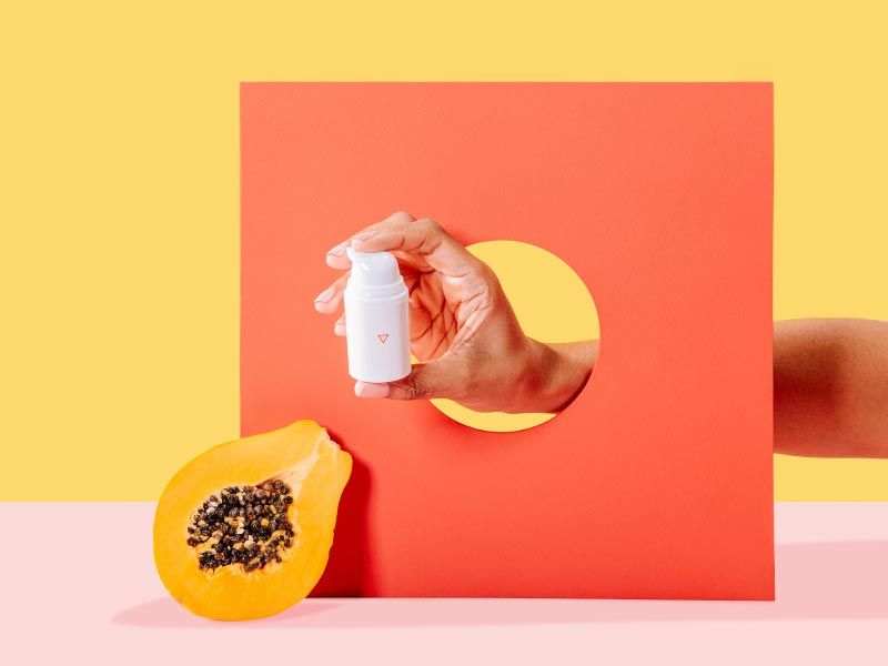 A person's hand holding a Wisp medication bottle through an orange cardboard cutout next to a cut open papaya with a yellow and pink background