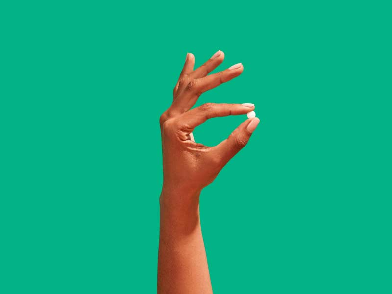 A woman's hand holding an emergency contraception pill in front of a green background