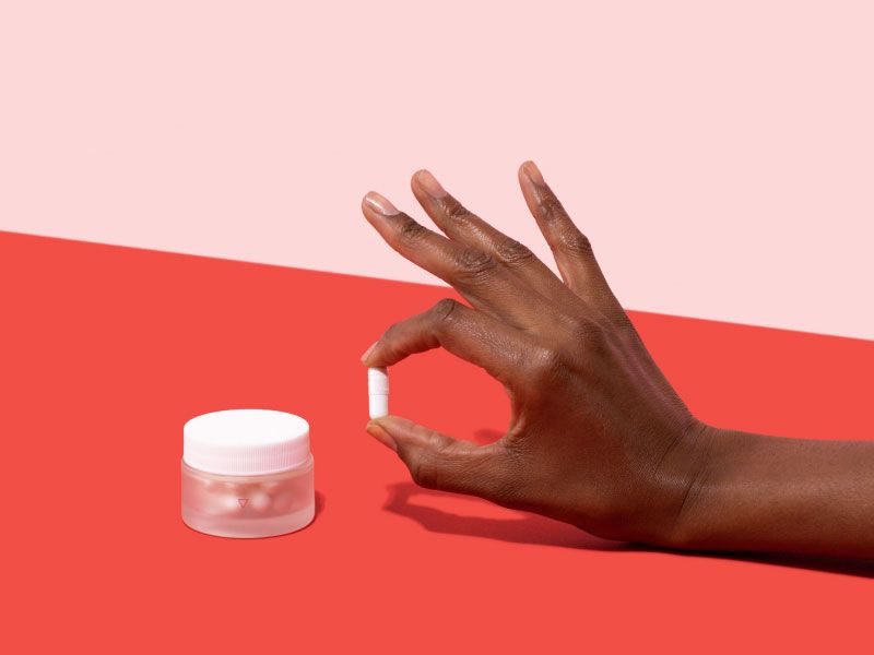 Woman's hand holding a Boric Acid pill next to a small glass Wisp jar on a pink and red background