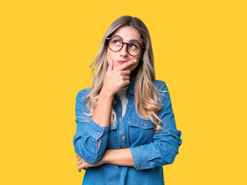 A woman with blonde hair and wearing a denim shirt and looking confused with a yellow background