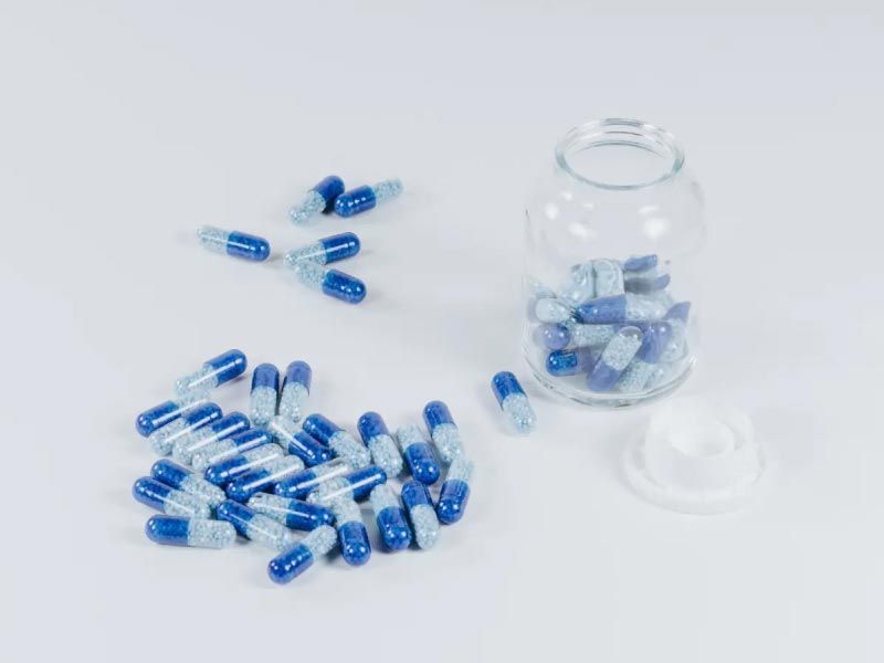 A clear pill bottle with blue and white pills inside and scattered around the bottle