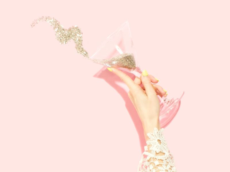 a woman's hand holding a martini glass pouring glitter onto a pink background