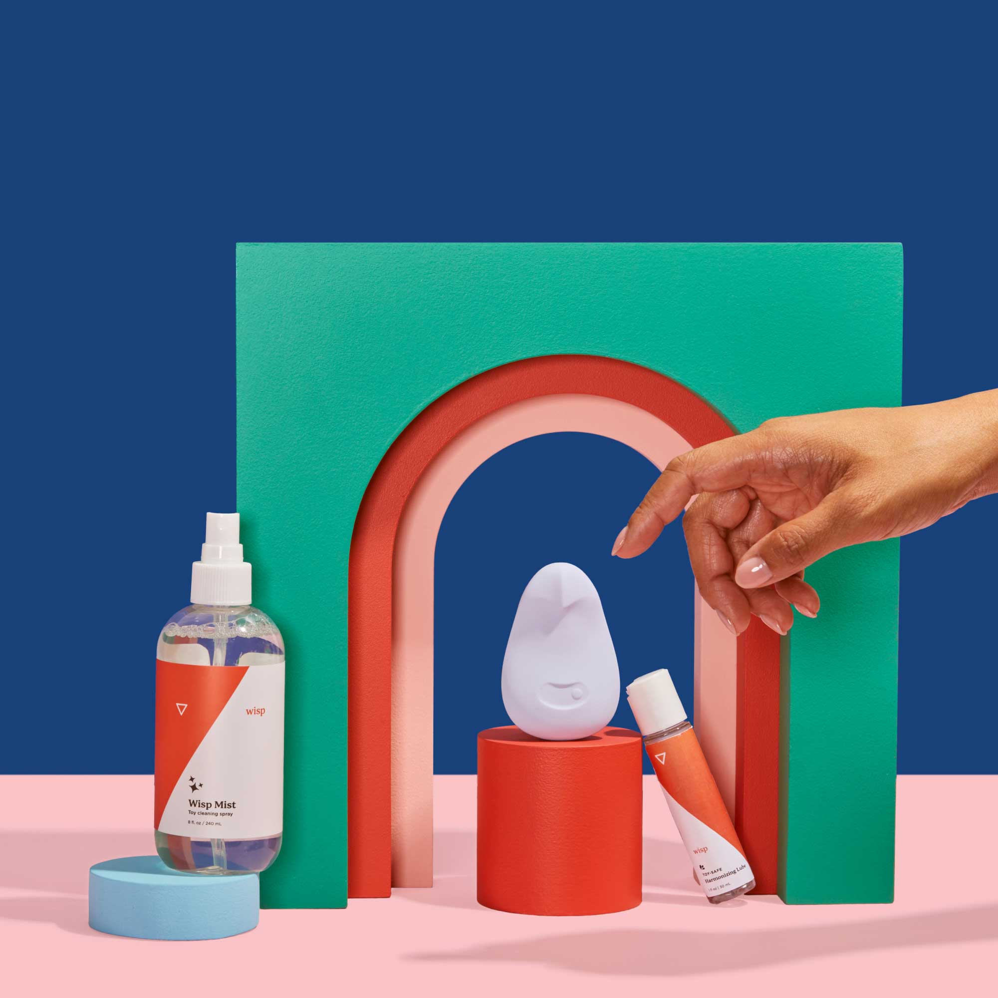 Hand reaching for Wisp Mist, Toy-Safe Lube, and Pom by Dame with colorful abstract shapes on a blue and pink background