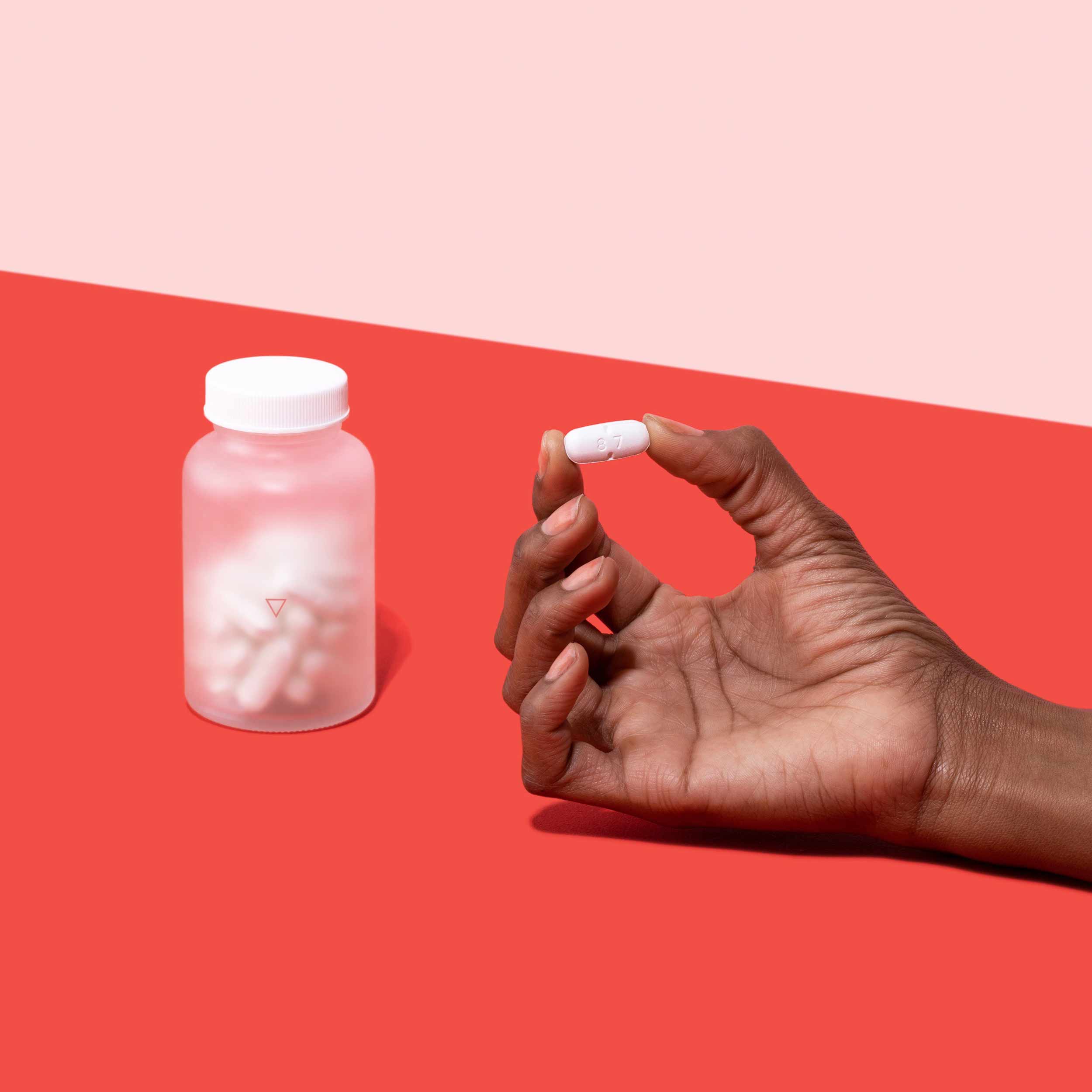Female hand holding Valacyclovir tablet next to bottle of Valacyclovir on a red surface, on a pink background