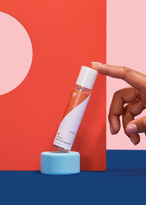 A female hand touching Wisp Original Harmonizing Lube with colorful abstract shapes on a blue surface with a pink background