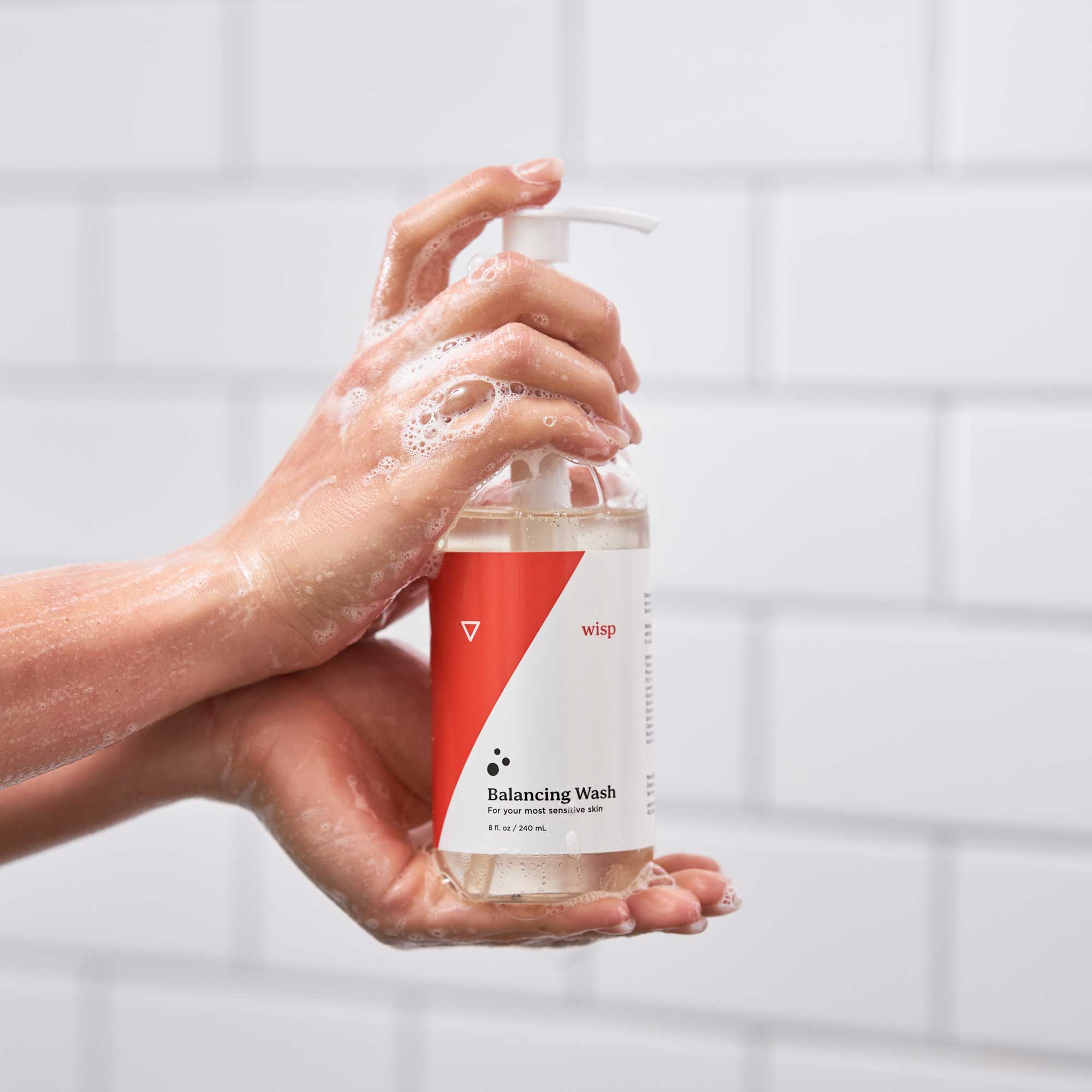Hands covered in soap suds holding a bottle of Wisp Balancing Wash against a white tile wall