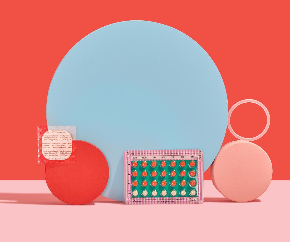 The birth control patch, ring, and pill are balanced on colorful abstract shapes on a pink surface with a red background