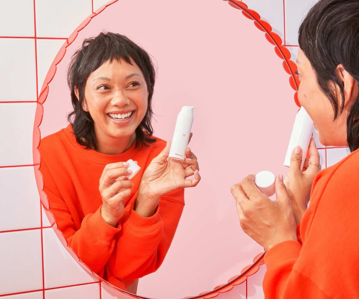 A woman wearing a red sweater is looking into a mirror and smiling while holding a Wisp skincare bottle