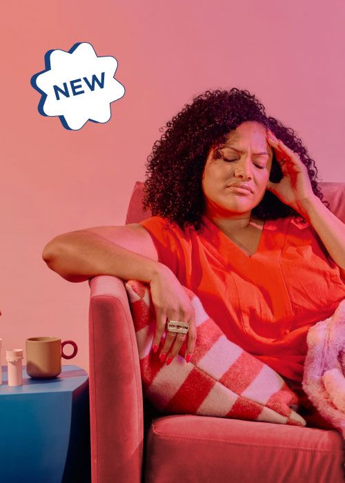 A woman wearing a red shirt it sitting on a pink sofa with her hand on her temple in obvious discomfort