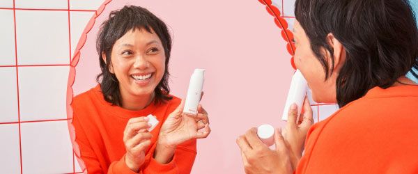 A woman wearing a red sweater is looking into a mirror and smiling while holding a Wisp skincare bottle