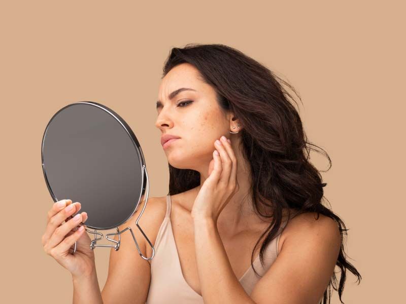 A woman touching her face and looking at her skin in a hand mirror