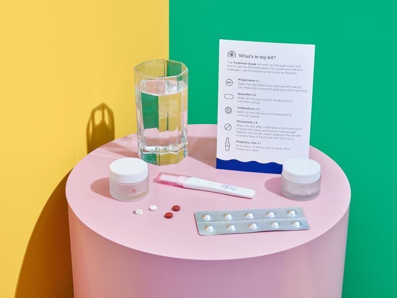 A pink nightstand with Wisp online abortion medications, treatment guide, and a glass of water, with yellow and green walls